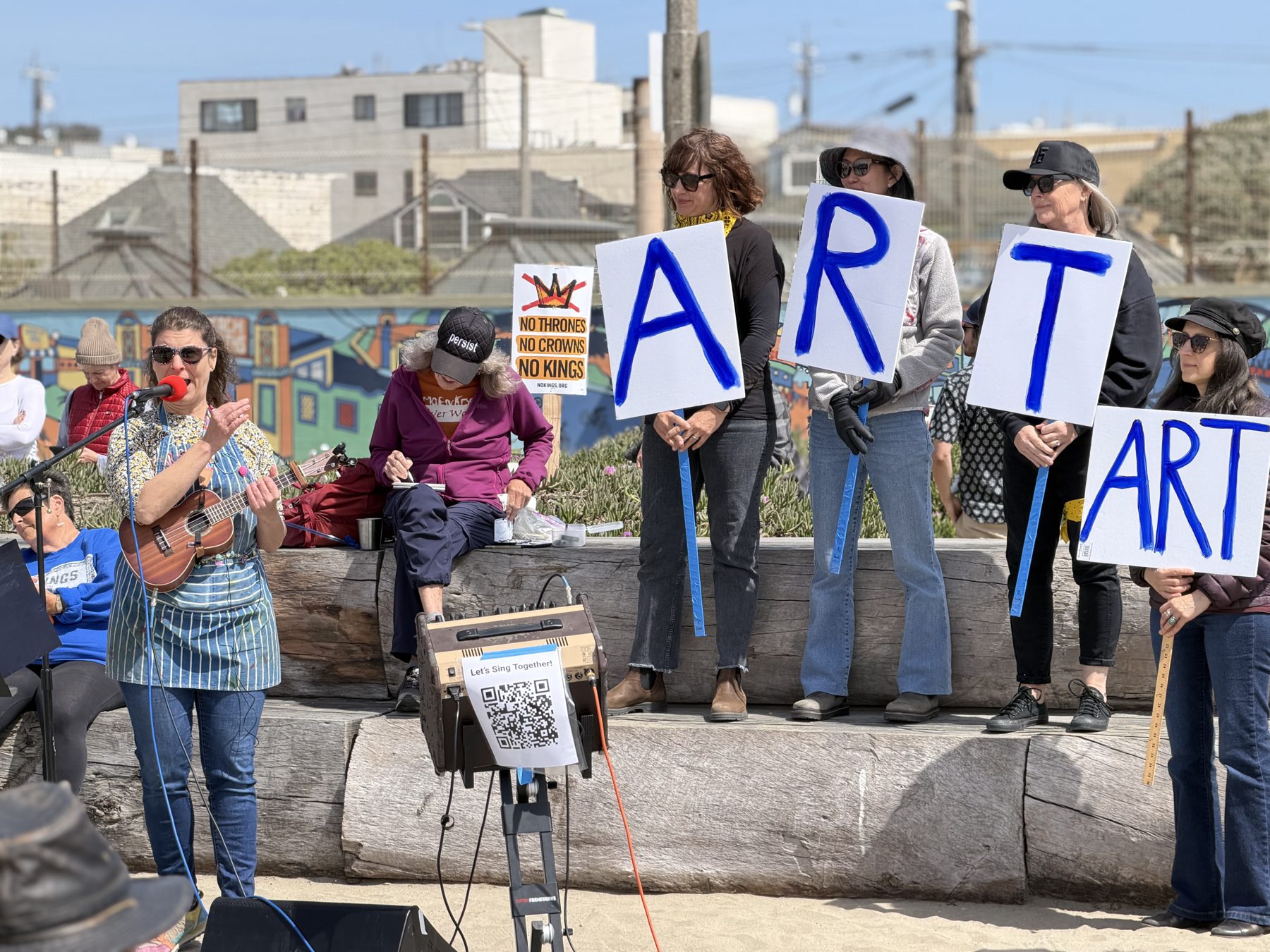 Volunteers holding hand-painted signs beside a singer with a ukulele at a westside rally